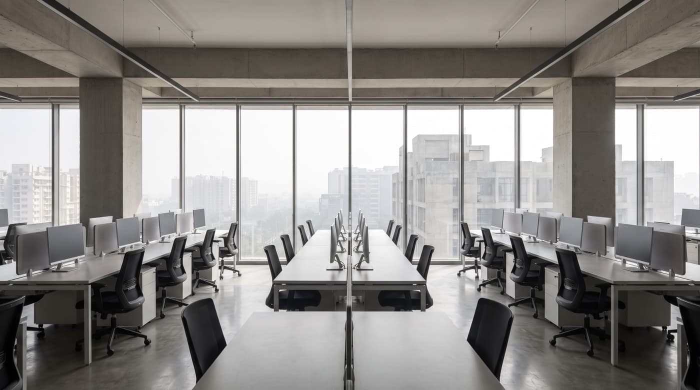 An empty contemporary open-plan office with rows of desks and monitors between bare concrete columns, looking out through full-height glazing onto a hazy city skyline.